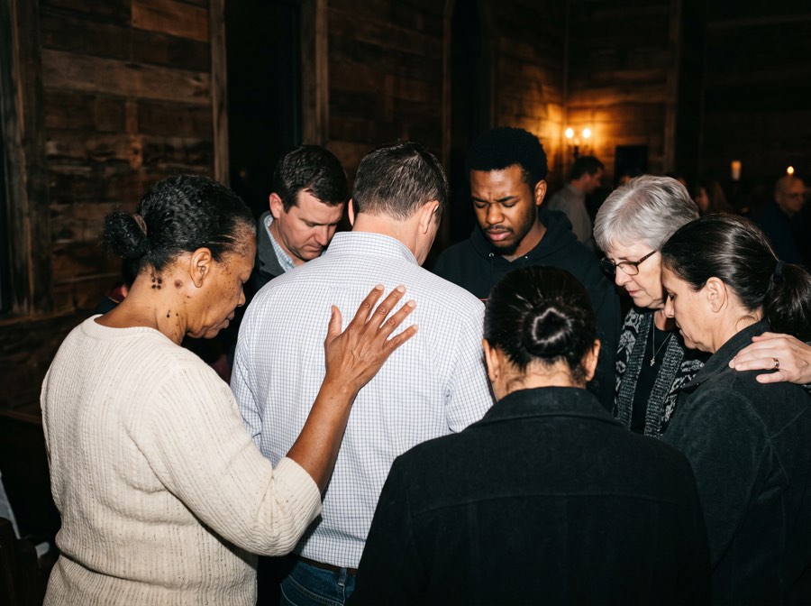 A small group standing in a circle with hands on one another, praying together.